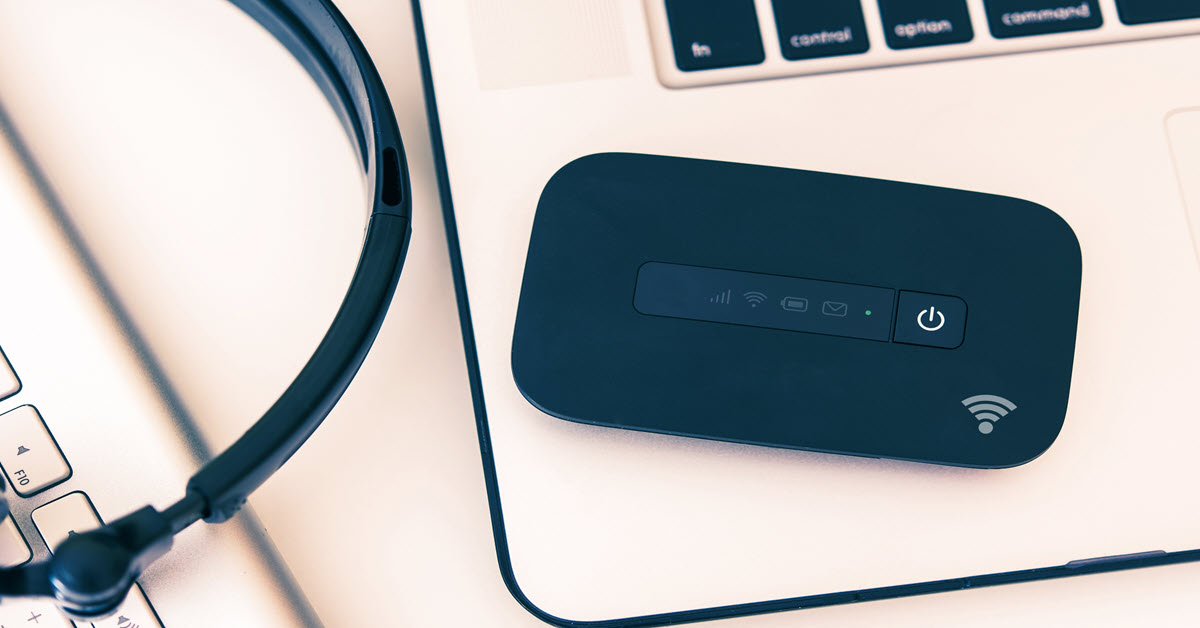 A laptop, mobile hotspot, and headphones sitting on a desk, representing portable internet access and remote work tools.